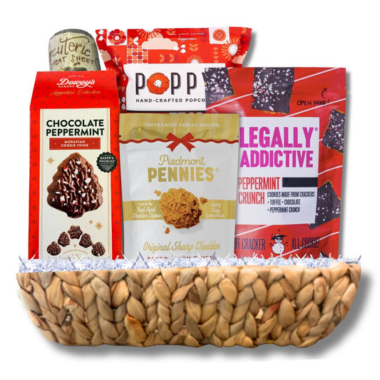 A snack and candy gift basket arranged in a woven natural-fiber bowl. Visible items include a box of Dewey's Chocolate Peppermint Cookies, a bag of Poppy Hand-Crafted Popcorn, a bag of Legally Addictive Peppermint Crunches, a box of cheese wafers and a wreath charcuterie mat.