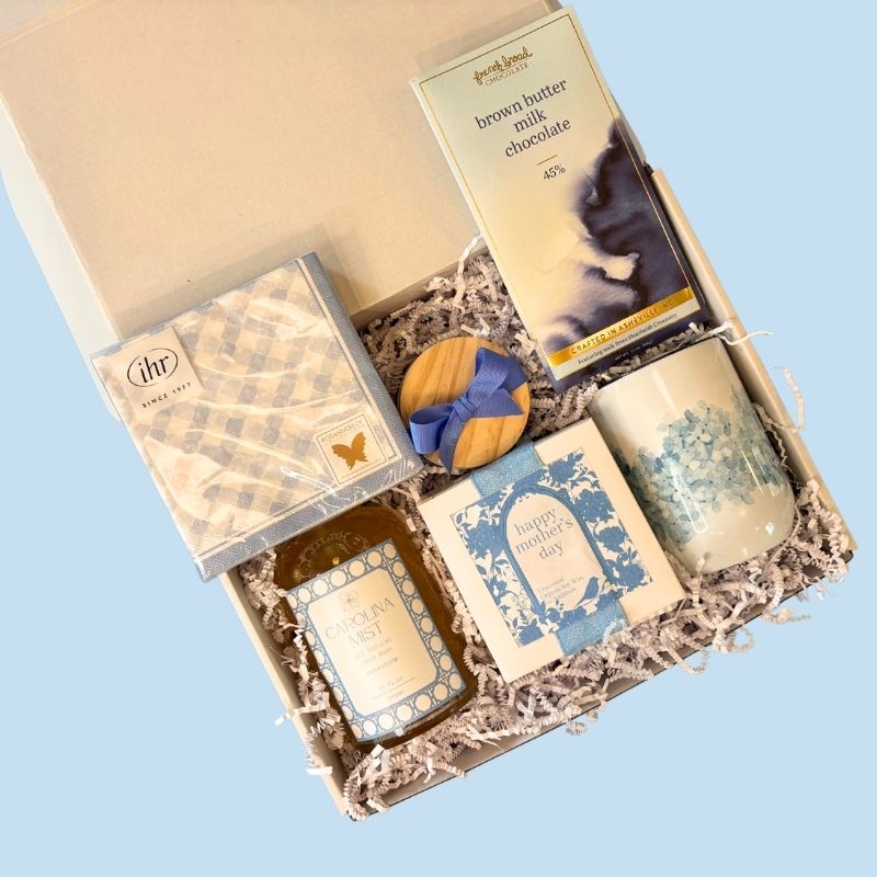 Top-down view of the luxury Mother's Day gift box showcasing NC-made treats, hand soap, soy candle, hydrangea tumbler, and gingham napkins arranged neatly inside.