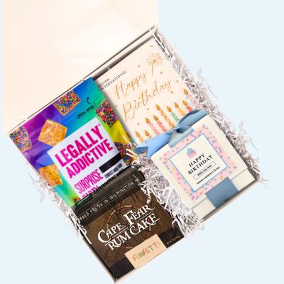 Overhead view of luxury birthday gift box open with North Carolina-made toffee cookies, rum cake, napkins, and candle arranged inside