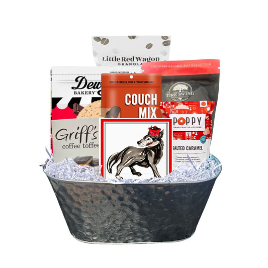 Assembled NC State-themed gourmet gift basket in a hammered tin planter, featuring red-packaged North Carolina-made snacks and watercolor wolf beverage napkins