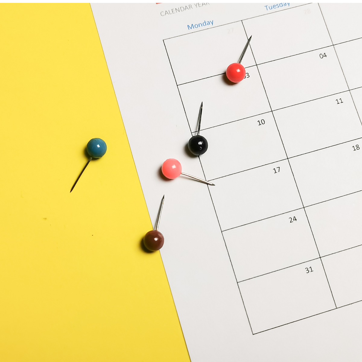 Five colorful pushpins with round beads are arranged on a white calendar page next to a bright yellow background.