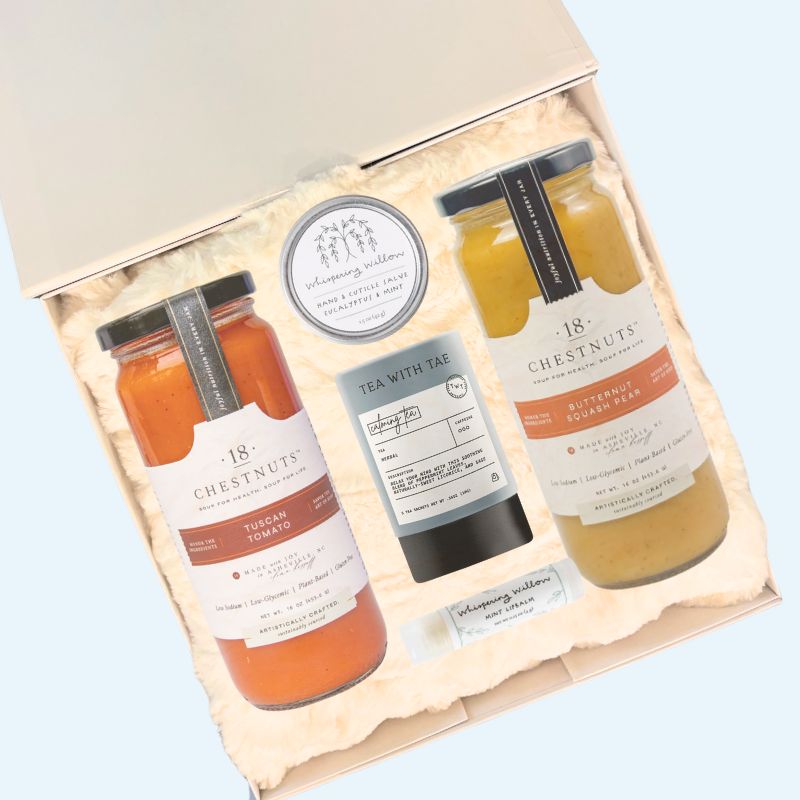 Open white luxury gift box with contents displayed, including a folded white faux fur lap blanket, two jars of 18 Chestnuts soup, Whispering Willow mint lip balm and eucalyptus hand salve, and a calming herbal tea sachet from Tea with Tae. Ideal for comfort, sympathy, or get well gifts.
