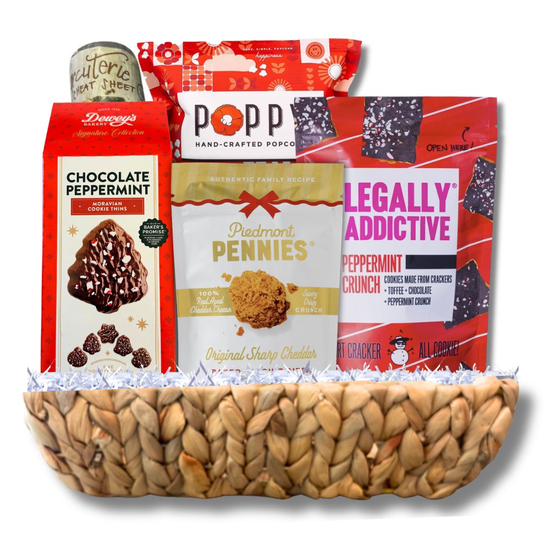 A snack and candy gift basket arranged in a woven natural-fiber bowl. Visible items include a box of Dewey's Chocolate Peppermint Cookies, a bag of Poppy Hand-Crafted Popcorn, a bag of Legally Addictive Peppermint Crunches, a box of cheese wafers and a wreath charcuterie mat.