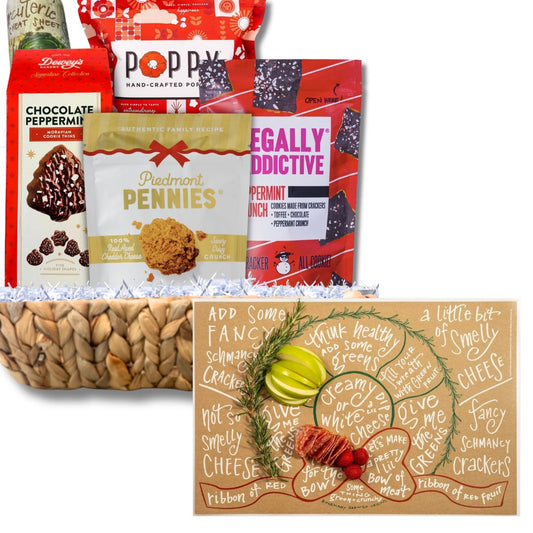 A gift basket filled with assorted packaged snacks and a decorative charcuterie mat. Visible products include bags of Poppy's Popcorn, Dewey's Chocolate Peppermint, Piedmont Pennies Cheese wafers, and Legally Addictive Peppermint Crunch crackers, arranged in white paper shred above a woven basket and a tan gift card with illustrated crackers and greenery.