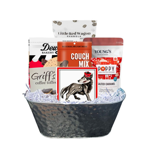 Assembled NC State-themed gourmet gift basket in a hammered tin planter, featuring red-packaged North Carolina-made snacks and watercolor wolf beverage napkin